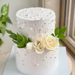 A classic two-tier wedding cake with a clean, modern aesthetic. The smooth buttercream is decorated with scattered pearls and a simple arrangement of fresh white roses and green leaves.