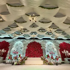 A regal reception stage where a canopy of white fabric and greenery drapes over a stunning backdrop of red roses, creating a look of classic opulence and romance.