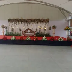 This is a full view of an elegant and simple stage design. It features a classic carved sofa for the couple, a backdrop of white drapes with hanging lights, and floral arrangements in large urns, all set on a red carpeted stage.