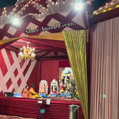 A traditional stage setup for a religious ceremony, draped in auspicious red and white fabrics and illuminated by a central chandelier.
