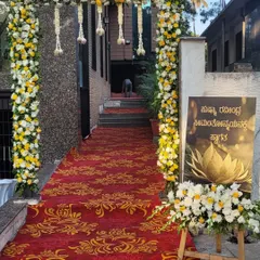 I create welcoming entrances that set the tone for the event. This one features a grand floral arch of white and yellow flowers over a red carpet, with a custom welcome sign in Kannada to greet the guests.