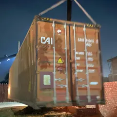 A crane carefully lifts the raw shipping container, positioning it for placement on the project site. This is the first step in giving the industrial object a new life.