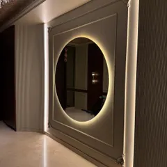 A stunning entrance foyer featuring a large, round mirror with warm backlighting. The design uses wall molding and vertical strip lights to create a grand and elegant first impression.