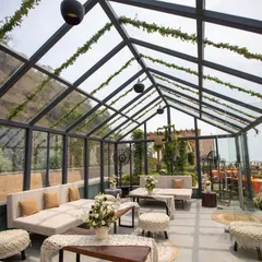 A beautiful glasshouse seating area, decorated with white sofas, macrame-topped stools, and natural-textured runners. The space is filled with light and greenery, creating a serene and rustic feel.