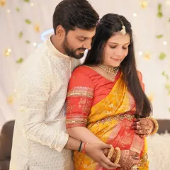 A beautiful candid shot of a couple at their baby shower. The father-to-be holds his wife from behind as they both look down at her belly, a moment of shared anticipation.