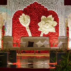 A stunning stage backdrop for a 'Ruby Romance' themed wedding. A wall of red roses is accented with white floral lotuses and framed by intricate, traditional white panels.