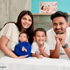 A happy family of four, including a newborn and a smiling older brother, posing comfortably on a bed for a relaxed, in-home style lifestyle photoshoot.