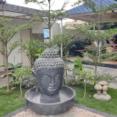 A large Buddha head sculpture converted into a water fountain, placed in the corner of a garden. The water flows gently from the top into the basin below.