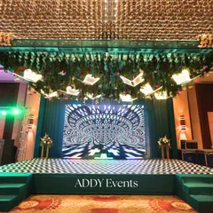 A stage designed for an unforgettable performance. The setup includes a massive LED screen with hypnotic visuals, a chevron dance floor, and unique butterfly-shaped lighting fixtures hanging from a lush green canopy.