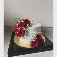 A simple yet dramatic wedding cake. The clean white buttercream provides a perfect canvas for a crescent of vibrant red roses, baby's breath, and gold accents, presented in a sleek black display box.