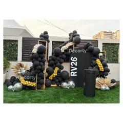 A wide shot of the outdoor photo backdrop for the 'RV26' birthday party. The asymmetric balloon arrangement in black, white, and gold creates a sophisticated and celebratory look.