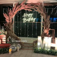 I design chic and trendy setups for any occasion. This rustic birthday decoration features a beautiful pampas grass wreath, a 'Happy Birthday' neon sign, marquee letter lights, and cozy, warm elements for a sophisticated celebration.