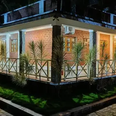 The "Grid in the Grove" farmhouse at night. The lighting design creates a dramatic and inviting ambiance, highlighting the rustic texture of the laterite block walls and the traditional form of the building.