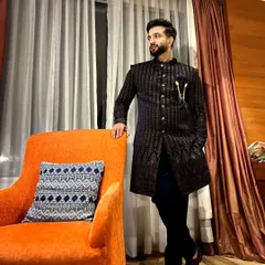 A full-length view of a dark, pinstriped Indo-Western sherwani. The outfit is accessorized with a stylish brooch, showcasing a modern approach to traditional groomswear.