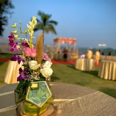 The same floral centerpiece seen during the day. The natural light highlights the fresh colors of the pink roses, purple orchids, and white tuberoses, with the outdoor wedding mandap visible in the background.