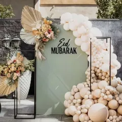 A modern and chic "Eid Mubarak" photo booth, with a sage green backdrop, a neutral-toned balloon arch, and dried floral arrangements.