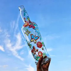 A hand-painted bottle with a floral and butterfly design, held up against the blue sky. The transparency of the glass makes the colors glow beautifully in the natural light.