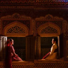 A cinematic shot of a couple in traditional attire, framed by the ornate windows of a palace. This style of photography uses architectural elements and dramatic lighting to create a regal and timeless portrait.