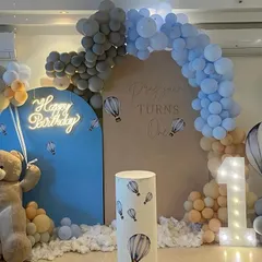 A modern and chic hot air balloon and teddy bear themed party. The decoration uses a soft color palette of grey, blue, and peach, with a neon sign and marquee number '1' for a trendy look.