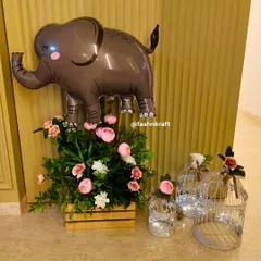 A cute and simple decor element for a baby girl's welcome party. An elephant foil balloon floats above a wooden crate filled with pink and white flowers, complemented by birdcages with fairy lights.