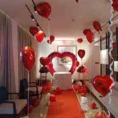 We transformed this room for a romantic birthday celebration. The decor includes a red carpet walkway, a heart-shaped arch made of red roses, and dozens of red heart-shaped foil balloons filling the space.
