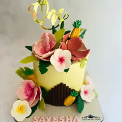 Celebrating a first birthday with a vibrant tropical-themed cake. This detailed creation features handcrafted sugar hibiscus flowers, plumeria, and miniature pineapples, all surrounding a custom gold acrylic "One" topper.