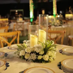 An elegant centerpiece perfect for a classic wedding theme. A lush arrangement of white carnations and chrysanthemums surrounds two pillar candles in glass hurricanes, creating a timeless and romantic look.