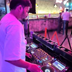 Bathed in pink light, I'm mixing a set in a busy club. I use my Pioneer controller to blend tracks smoothly, creating a continuous flow of dance music that builds throughout the night.