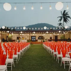 The guest seating for a Sangeet under the stars. The vibrant orange chair covers add a pop of color, while strings of paper lanterns and bulbs create a festive and magical ambiance against the twilight sky.