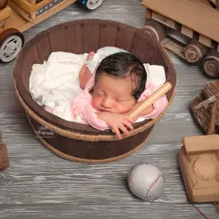 This posed newborn shot has a rustic, playful theme. The baby is nestled in a wooden barrel, surrounded by charming wooden toys, a miniature baseball bat, and logs, creating a unique and cozy portrait.