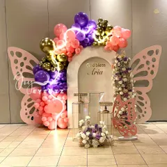 A vibrant butterfly-themed decoration for a First Holy Communion. This design features large pink butterfly wings, a colorful balloon arch with neon light accents, and clear plinths for display.