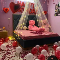 A full view of the birthday room setup, with "HAPPY BIRTHDAY" foil balloons, a light-up canopy, teddy bears, and a floor covered in balloons and petals.