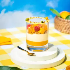A layered mango parfait in a glass, presented on a white pedestal against a sunny, picnic-themed background. The bright colors and clean composition make it look refreshing and delicious.