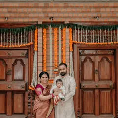 A classic family portrait of the new parents with their baby, taken at their Bangalore home. The traditional wooden doors and marigold decorations create a perfect backdrop for a timeless family photograph.