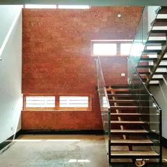 This is an interior view of a courtyard in a Bengaluru residence. The feature wall is clad in laterite stone, adding a warm, earthy texture that contrasts beautifully with the sleek glass and metal staircase.