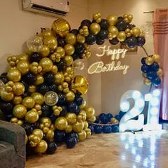 Celebrating a 21st birthday in style. This setup features a dramatic black and gold organic balloon arch, a custom 'Happy Birthday' neon sign, and large, illuminated marquee numbers to mark the special occasion in any home.