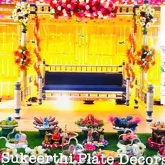 A vibrant baby shower setup at Brookefields, Coimbatore, with a beautiful swing as the centerpiece. The 15 platters on the grass floor are filled with colorful sweets, fruits, and thematic dolls.