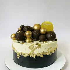 A sophisticated design combining black, white, and gold. The bottom half is coated in smooth black buttercream, while the top features a fault-line edge with gold leaf and an assortment of handcrafted chocolate spheres in both matte and shiny gold finishes.