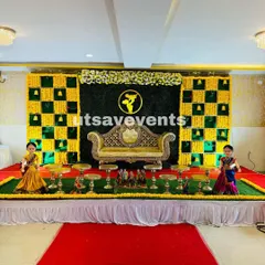 A wide view of a classic seemantham stage decoration in a convention hall. The design features a green and yellow checkerboard floral backdrop, a golden Diwan sofa, and a symmetrical arrangement of plate stands on a red carpet.