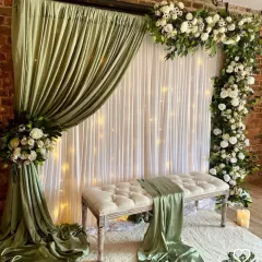A very elegant and unique backdrop idea for a reception or engagement. This setup uses sage green drapes, an asymmetrical white floral arrangement, and a tufted bench for a soft, romantic feel.