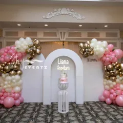 A beautiful stage for a girl's First Communion, featuring pink, white, and gold balloon arrangements. The central white arch backdrop includes a custom name cutout and a pedestal for the cake.