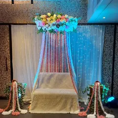 A canopy-style seating arrangement for a Mehndi function. This design features a floral chandelier with hanging flower strings in pink and white over the main seat.
