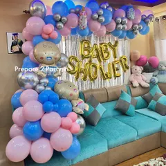 A classic at-home baby shower decoration. The setup features a pink and blue balloon arch over a couch, a silver shimmer curtain, and large gold foil letters spelling "BABY SHOWER".