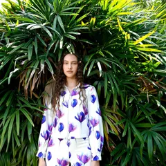 This look pairs a white and purple floral print shirt with matching trousers. The lush green foliage in the background makes the purple hues of the outfit pop.