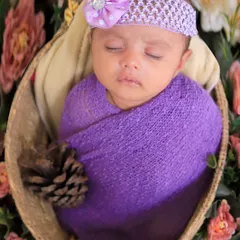 This newborn is wrapped in a vibrant purple swaddle and placed in a hanging basket prop. The floral background creates a dreamy, garden-like setting for the portrait.