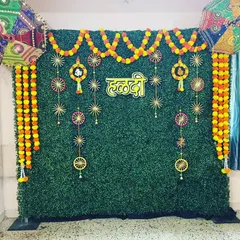 A beautiful Haldi decoration perfect for home celebrations. An artificial green wall is adorned with marigold garlands, a 'हळदी' sign, and colorful Rajasthani umbrellas, creating a perfect photo booth.