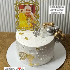A cake made with devotion for a Guruji celebration. This elegant white cake is decorated with a photo of Guruji in a golden frame, delicate butterflies, and gold and silver spheres.