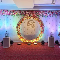 A beautiful and modern stage setup for an engagement ceremony. The centerpiece is a large circular ring completely covered in multi-colored flowers, with the couple's initials in the center, creating a perfect photo backdrop for the special day.