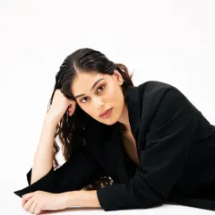 A striking high-fashion portrait against a clean white background. The model's pose and direct gaze, combined with the simple black blazer, create a minimalist yet powerful editorial image.