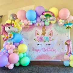 An enchanting unicorn-themed first birthday party. A custom backdrop is framed by a pastel rainbow balloon garland, complete with a large unicorn foil balloon and a number "1" balloon.
